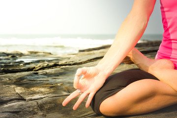 Fototapeta na wymiar Female yogi hand practicing meditation on the rocks by the sea at sunrise. Yoga teacher in padmasana on the beach. Instructor on lotus pose, chin mudra, peaceful mindfulness concept