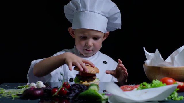 Time lapse of cheerful boy cooking a burger. He wears chefs suit and cap. Black background