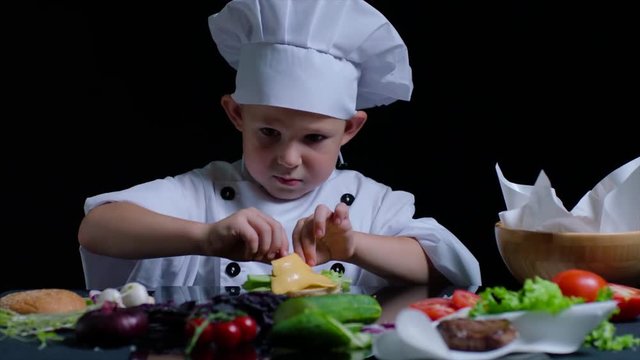 Time lapse of cheerful boy cooking a burger. He wears chefs suit and cap. Black background