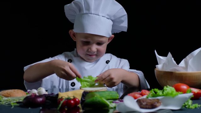 Pleasant boy cooks a burger, wearing chefs suit and cap. Black background for commercial