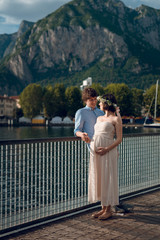 Portrait of happy young couple of pregnant woman and her husband. Man hugging his wife while standing near the lake Como, Italy with scenic view of rocky mountain.