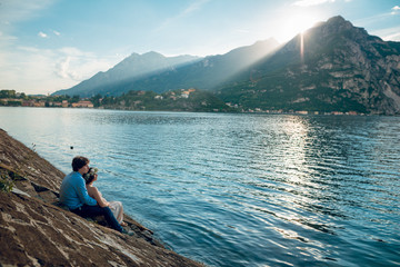 Fototapeta na wymiar Young couple of pregnant woman and man sitting on the stairs on the shore of Lake Como with wonderful mountain view on sunny summer day