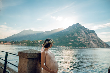 Fototapeta na wymiar Beautiful young pregnant woman in tender dress and flower wreath looking at the scenic view of lake Como and mountains on background. Sunny summer day in Italy. Pregnancy