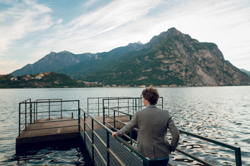 Rear view of young man standing on the pier on the lake Como with scenic view of beautiful mountains. Summer day. Italy