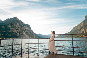 Fototapeta na wymiar Young tender pregnant woman in nice dress standing on the pier near the Como lake. Scenic view beautiful mountains and lake. Summer day