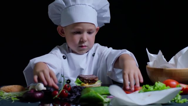 Pleasant boy cooks a burger, wearing chefs suit and cap. Black background for commercial