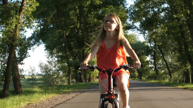 Cute blonde teenage girl in glasses on a bicycle trip in summer countryside. Sweet teen girl enjoying views of nature while riding a bike on rural road. Steadicam shot. Front view