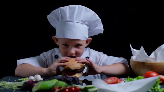 Cute boy is eating a burger while cooking. He wears chefs suit and cap.