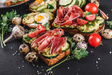 Various of sandwiches and bruschetta with prosciutto, fried quail egg, avocado, cucumber, tomatoes, on black stone background.