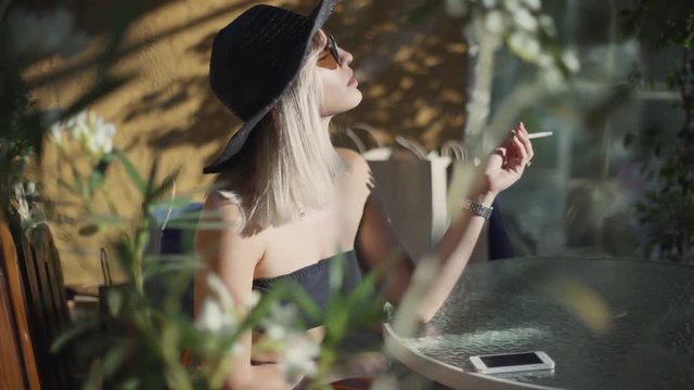 Beautyful stylish young blonde woman smoking cigarettes outdoors in han and sunglasses