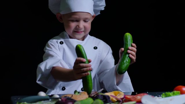Cute boy wearing chefs suit plays with cucumbers at the cooking table. Black background