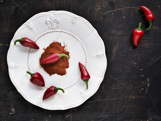 Fresh chilli peppers and pepper powder on white plate and dark table. Flat lay.