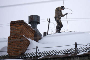 a young man in black dresses is standing on a red roof and cleaning the chimney with a metal brush on a long cable.