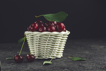 Fototapeta na wymiar White basket with ripe cherries and green leaves on a black background.