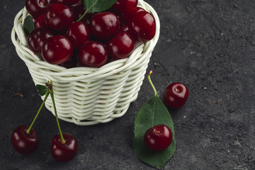 White basket with ripe cherries and green leaves on a black background. Copy space