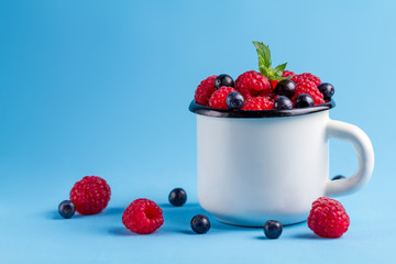 Raspberries, blueberries and a sheet of fresh mint in a white cup on a blue background.