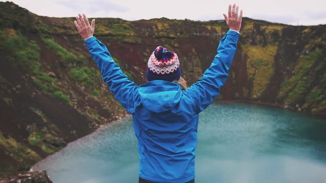 Young tourist woman in jacket and hat rise her hands enjoying beautiful view of Kerid lake of Iceland
