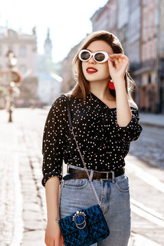 Outdoor portrait of yong beautiful happy smiling woman wearing stylish sunglasses, black polka dot blouse, blue mom jeans, with small quilted bag. Model posin in street of european city. 
