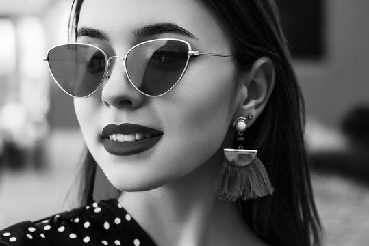 Outdoor close up monochrome portrait of young beautiful stylish woman wearing cat eye sunglasses, tassel earrings, posing in street. Female fashion concept