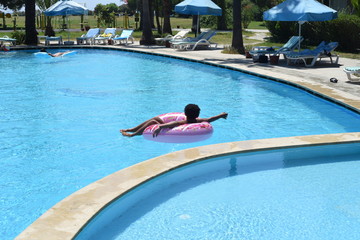 Happy cute little african-american boy lying on an inflatable donut circle  in the swimming pool in the hotel. Cool summer holidays for children and kids. Active games on the water.  Pink donat. 
