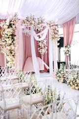 Fototapeta premium The wedding ceremony area is decorated with white and peach cloth, crystal chandelier, transparent chairs for guests and beautiful floral arrangements of roses