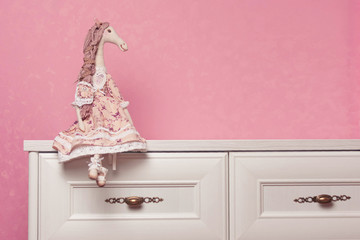 The handmade doll sits on a chest of drawers against the backdrop of a pink wall. A doll is a horse in a dress. Puppet in the interior.
