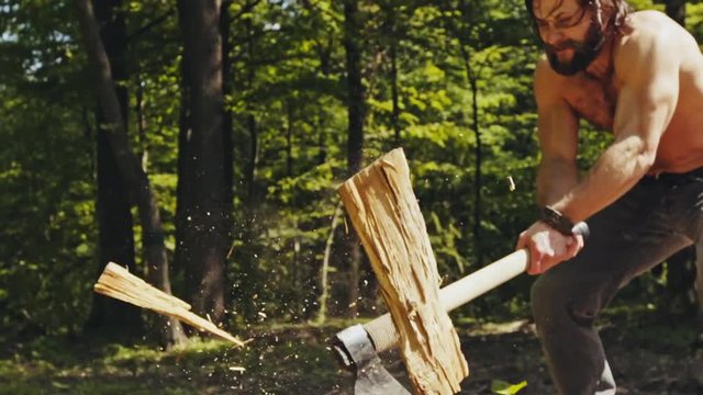 Strong shirtless man splitting logs in woodland
