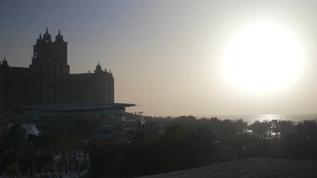 huge sun disc is hanging over sea surface, palms and amazing buildings
