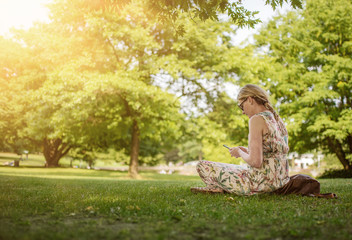 low angle view of woman sitting cross-legged on lawn in public park using her smartphone on sunny summer day