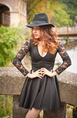 Charming young light brown hair brunette woman with big black hat and blouse with lace sleeves. Sexy gorgeous young woman with curly hair posing. Beautiful portrait of a sensual woman with long hair