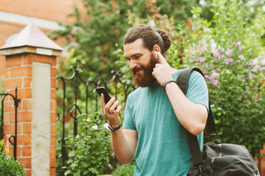 Cheerful happy bearded man looking at his smartphone, listening to the music with earpods outdoors.