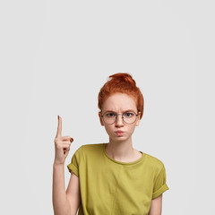 Angry female with ginger hair combed in bun, points upwards with fore finger, shows blank space for...