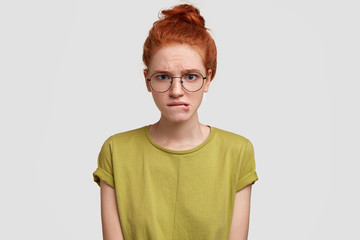 Nervous ginger female bites lips, has discontent expression, doubts about something, wears casual t shirt, isolated over white studio background. Unhappy upset foxy woman can`t make decision