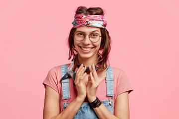 Happy female hippy makes intriguing gesture, looks with cunning expression, smiles gently, has mysterious plan in mind, raises eyebrows, dressed in stylish overalls and headband, stands alone indoor