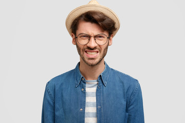 Horizontal shot of irritated unshaven male clenches teeth, frowns face, wears straw hat and denim shirt, feels annoyance, isolated over white background. People, emotions and reaction concept