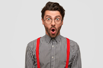 Astonished young European male with terrific expression, keeps mouth opened, stares with bugged eyes, can`t believe in shocking news, isolated over white background. People, terror, fear concept