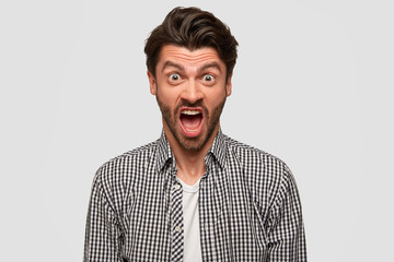 Aggressive furious bearded male has annoyed facial expression, raises eyebrows, shouts at someone during quarrel, argues in irritation, has dark hair, isolated over white background, dressed in shirt