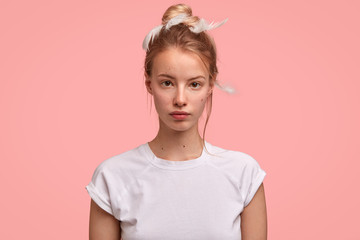 Photo of serious lovely Caucasian female looks directly at camera, has sleepy expression, wears casual t shirt and feathers, can`t gather with thoughts early in morning, isolated over pink background