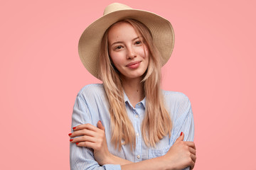 Beautiful pleased shy young female model keeps hands crossed, looks positively at camera, wears hat and shirt, isolated over pink background. Fashionable European woman tourist has excursion