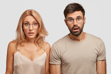 Two emotive stunned young female and man in nightwear, overslept train, stare at camera with puzzled shocked facial expressions, stand next to each other, isolated over white studio background.