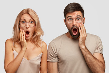 Studio shot of shocked wife and husband find out they will become grandparents very early, keep jaws dropped, stand closely together against white background. People, reaction, relationship concept