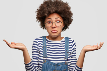 Clueless African American female with curly bushy hair, shrugs shoulders, looks doubtfully as faces dilemma, wears striped sweater and jean overalls, stands against white background. People and doubt - obrazy, fototapety, plakaty