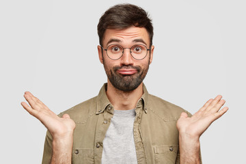 Photo of puzzled frustrated hesitant bearded male presses lips together, shrugs shoulders, makes...