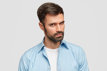 Confident unshaven guy with dark bristle and hair, listens attentively boss, stands against white background wears blue shirt. Serious male entrepreneur decides on new plan. Facial expressions concept