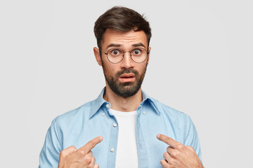 Studio shot of indignant bearded male points at himself, being shocked by something, shows blank space on clothes for your logo or design, isolated over white background. People and emotions