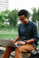 Fototapeta premium Young man university student online learning via laptop computer, sitting on a bench on a campus. Stylish hipster guy experienced freelancer working distance on notebook while resting in city park
