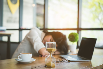 Freelancer asian businesswoman tired after working coffee shop her sleeping on workplace table near windows and hourglass at evening with digital laptop computer and coffee break.