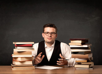 A young teacher in glasses sitting at classroom desk with pile of books in front of clean blackboard back to school concept.