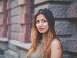 Obraz na płótnie Canvas Portrait of young attractive girl looks into the camera with smile. Woman with oriental face, brown eyes and stylish ombre dyed long haistyle on architecture background. Street style.