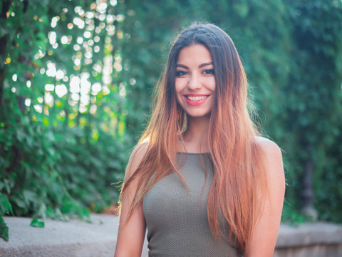 Portrait of young attractive girl looks into the camera with smile. Woman with oriental face, brown eyes and stylish ombre dyed long haistyle on foliage background. Street style.
